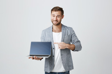 Stylish male enterpreneur with thick beard and trendy hairstyle posing against gray studio background with laptop in hands, smiles at camera, points at screen, demonstrates how good this device is.