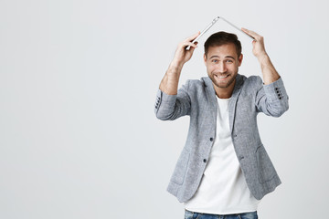 Positive young caucasian bearded man wearing trendy jacket over white t-shirt holding modern laptop computer over his head, smiling with teeth at camera, posing against gray background.