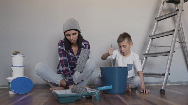 A young girl sits on the floor near the wall and holds two rollers in her hand, and her brother sits next to her and mixes the paint that is in a bucket mixer