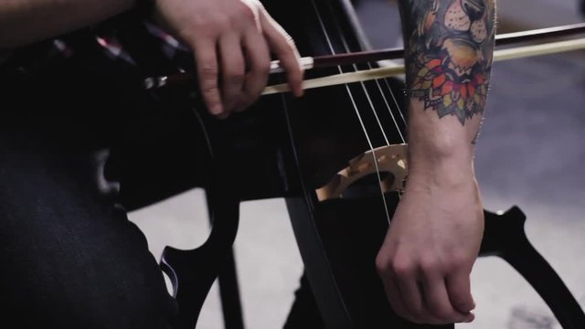 young man play on electric cello in studio, hipster musician with tattoo