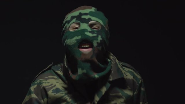Portrait of a laughing soldier in camouflage and a military mask standing over black background