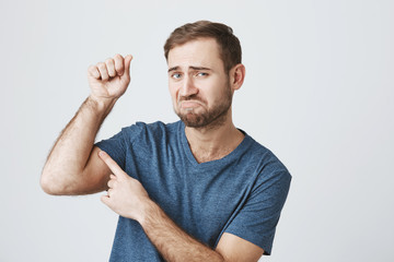 Funny dissatisfied bearded guy wearing casual clothes raising arm with pumped fist, tensing muscles to show his biceps and strength, curving lips, displeased with results of first training at gym