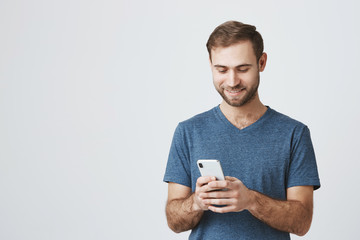 Happy glad pleased man with stubble smiling to the camera reading positive news or browsing newsfeed via social networks using smartphone, having small break, posing indoors.