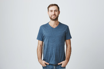 Fototapeta premium Handsome bearded man wears blue t-shirt and jeans has good mood, holds his hands in pockets, smiles at camera, poses indoors. Caucasian male model expresses positive emotions.