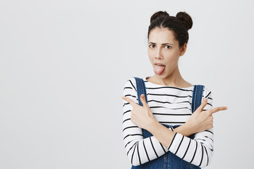 Young woman with two hairbuns looking at something with disgusting look pointing index fingers, sticking out tongue, showing copy space for advertisement. Funny girl in denim overall and striped top