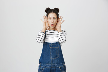 Playful positive carefree brunette girl with two hairbuns with opened mouth, has cheerful expression, wears denim overalls, has fun, holds opened palms behind ears, poses against studio background.