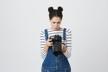 Puzzled emotional young pretty caucasian female photographer with two hairbuns wearing denim overall using modern camera, looking at pictures she has just taken, pouting her lips