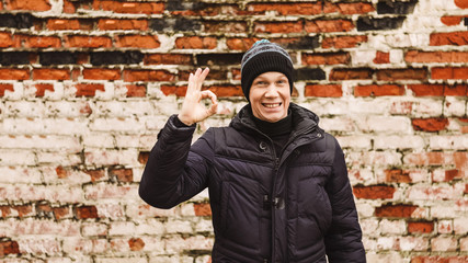 the guy shows the gesture ok. Brick wall . hat and jacket. smile on the face