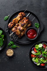 Grilled meat skewers, shish kebab and healthy vegetable salad of fresh tomato, cucumber, onion, spinach, lettuce and sesame on black background, top view
