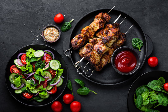 Grilled meat skewers, shish kebab and healthy vegetable salad of fresh tomato, cucumber, onion, spinach, lettuce and sesame on black background, top view