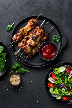 Grilled meat skewers, shish kebab and healthy vegetable salad of fresh tomato, cucumber, onion, spinach, lettuce and sesame on black background, top view