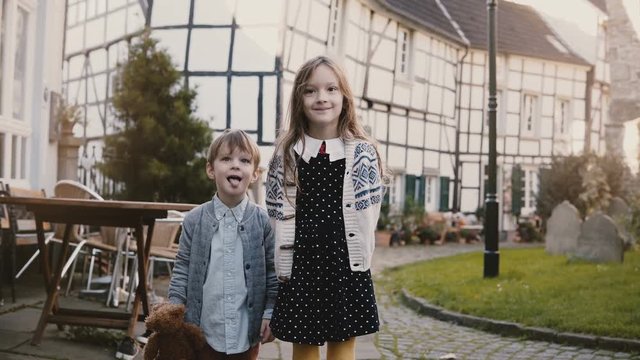 Little girl and boy stand together making faces. European siblings look at camera smiling silly. Half-timbered house 4K.