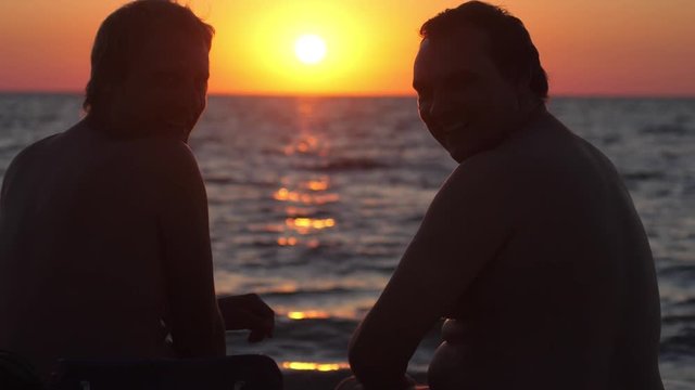 Young man sit back and relax at the beach in sunset time. Two gay friends flirt and drink beer. slow motion. 1920x1080