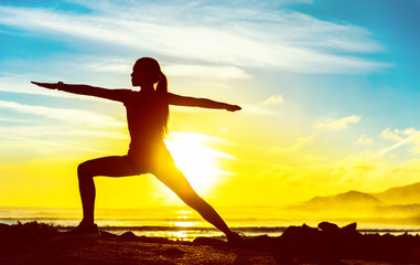 Yoga woman meditating zen in warrior pose relaxing outside by beach at sunrise or sunset. Female yoga instructor working out training in sun flare.