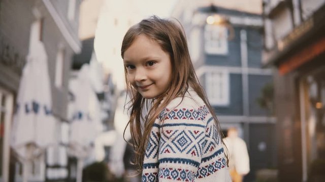 Portrait of little European girl 5-7 years old. Posing at camera smiling, amazing long hair. Old houses background. 4K.