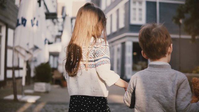 Camera follows little girl and boy walking together. Back view. Two kids wander around old town. Brother and sister. 4K.
