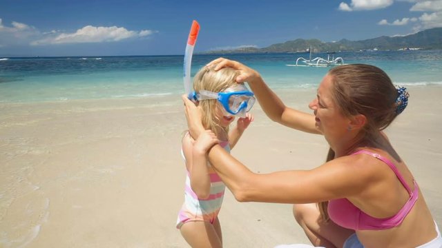 Caucasian mother helps little daughter to put on snorkel mask on white sand beach with clear water