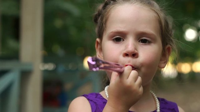 Happy little girl are blowing into unfolding tube at a birthday party. Little girl having fun and blows a party horn. Little girl in purple dress with birthday pipes.