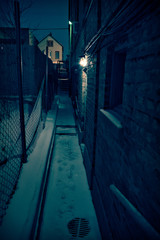 Dark and scary urban city gangway during the winter with snow and foot prints by a vintage brick building at night.