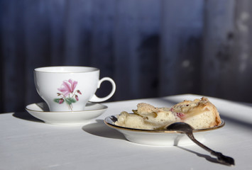 on the table a plate with a piece of apple pie, a spoon and a cup in the sunbeam 