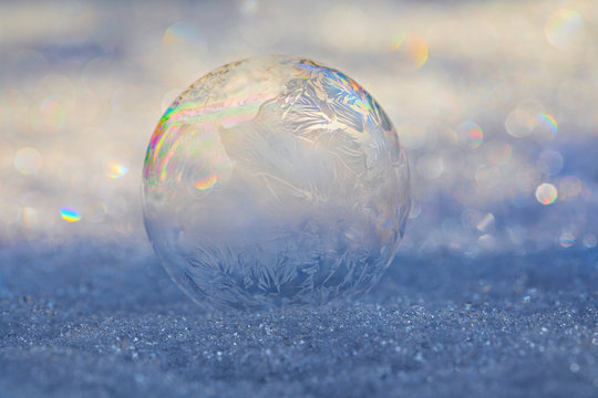 A frozen soap bubble on the snow with prism like highlights, ice crystals forming on the bubble, and bokeh from the sun on the snow