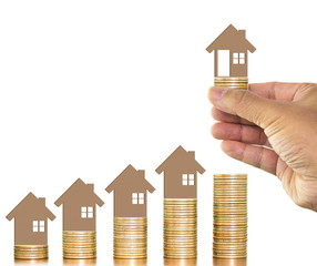 Man hand holding money coin with wooden toy house putting on money coins stack arranged as a graph on wooden table white background, concept of money growth and saving money