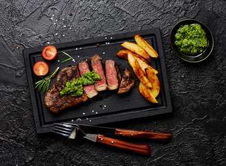 Meat Picanha steak, traditional Brazilian cut with potato wedges, chimichurri sauce and rosemary on black meat cutting board. Steak and a glass of red wine with fork and knife.