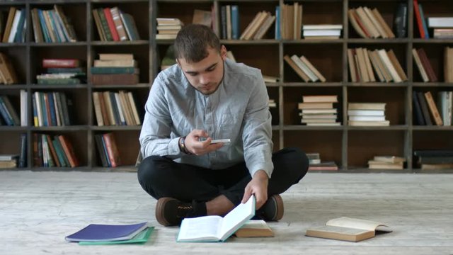 Carefree male student with smart phone taking picture of books pages while sitting cross-legged on the floor in campus library. Hipster man with mobile phone making cheat sheet in university library