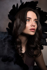 Vertical beauty portrait in dark tones. Beautiful young woman in a cloud of smoke with black feathers in her hair