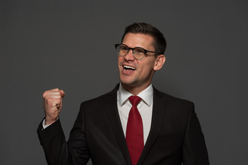 Portrait of a positive successful businessman wearing glasses and formal clothes showing yes and holding his hand up against gray background