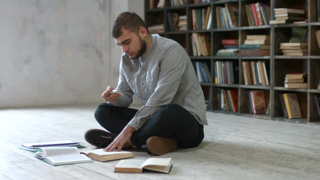 Dishonest male college student with mobile phone picturing books pages while sitting on the floor against bookshelf in campus library. Hipster with smartphone making cheat sheets for exam in library.