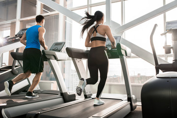 Full length portrait of two fit young people, man and woman, running on treadmills facing windows...