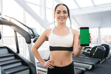 Fototapeta na wymiar Waist up portrait of fit young woman wearing sports clothes presenting mobile app holding smartphone with green screen and smiling at camera