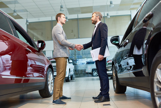 Full Length Portrait Of  Handsome Client Shaking Hands With Mature Sales Manager In Car Showroom, After Buying Brand New Luxury Car