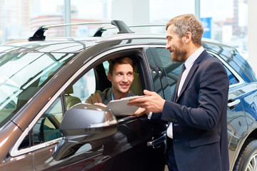Portrait of handsome young man taking luxury car for test drive, sitting inside and smiling at sales manager
