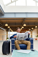 Fototapeta na wymiar Full length portrait of handsome young man with suitcase reading newspaper sitting in leather armchair while waiting for check in in modem hotel