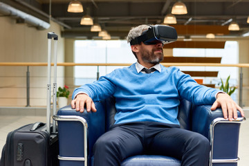 Fototapeta na wymiar Portrait of mature bearded man wearing VR headset sitting in armchair immersed in game and looking around, copy space