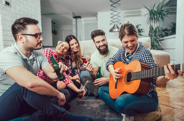 Group of happy young friends with guitar having fun and drinking beer in home interior