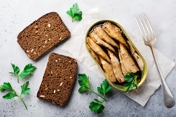 Canned sprats in tin can with fork with bread for making sandwich on gray old concrete or stone background. Selective focus. Rustic style. Top view. Copy space.