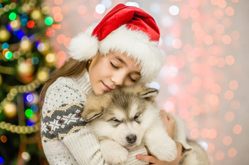 Fototapeta na wymiar happy little girl in red santa hat hugging puppy on a background of the Christmas tree