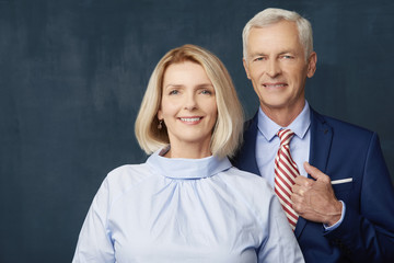 Senior couple portrait. Senior couple standing together at dark background. Beautiful blond woman looking at camera and smiling while elegant old man standing next to her. 