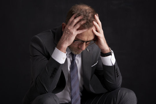 Stressful businessman portrait. Portrait shot of a stressful middle aged businessman wearing suit while sitting at dark background with hands on his forehead. 
