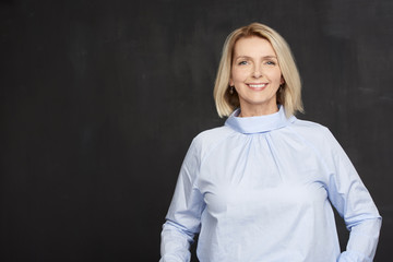 Fototapeta premium Confident blond female portrait. Close-up studio shot of a beautiful senior woman wearing shirt while standing at dark background and smiling. 