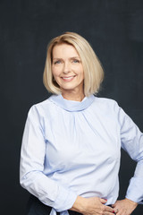 Confident blond female portrait. Close-up studio shot of a beautiful senior woman wearing shirt while standing at dark background and smiling. 