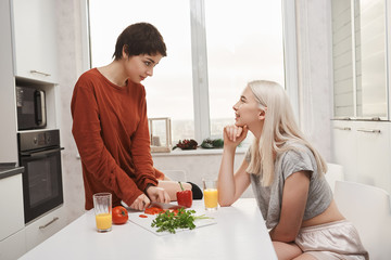 Portrait of two woman sitting in kitchen, drinking juice and making salad while talking and making...