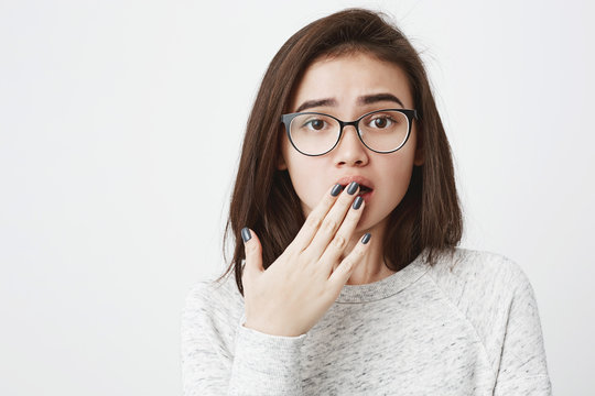 Attractive brunette european student in glasses, covering opened mouth with hands, expressing tiredness over white background. Girl hears her friend secret imitating she never heard it before.
