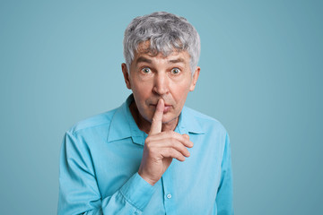 Horizontal shot of grey haired male looks with puzzled expression at camera, keeps fore finger on lips, asks to be quiet and mute, demands hush, dressed in elegant formal shirt isolated over blue wall