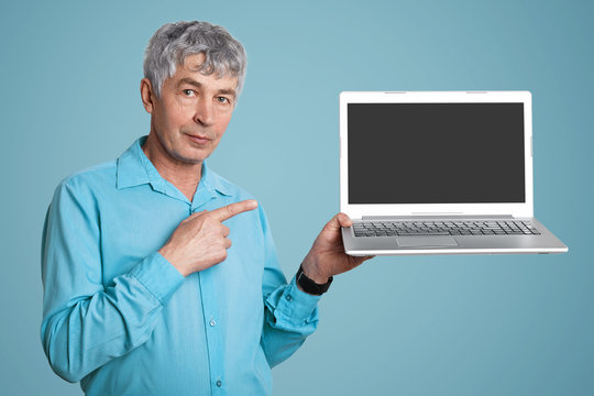 Elderly wrinked handsome male in formal shirt holds modern portable laptop computer with blank copy screen for your text or advertising content, indicates at gadet, isolated over blue background