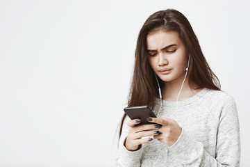 Attractive young female with worried expression holds phone and listen music, over white...