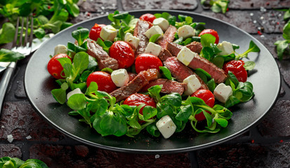 Juicy Beef Sirloin Steak Salad with roasted tomatoes, feta cheese and green vegetables in a black plate. healthy food